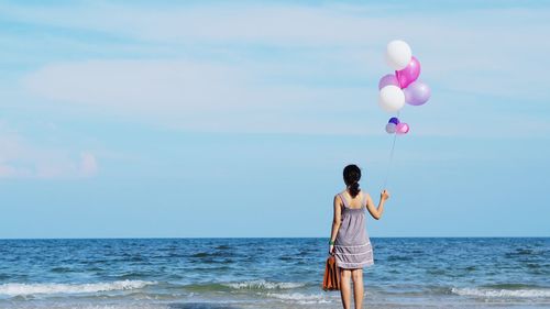 Rear view of woman with balloons on shore at beach against sky