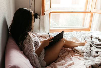 Woman writing in book while sitting on bed at home