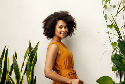 Side view portrait of young woman smiling while standing amidst plants against wall