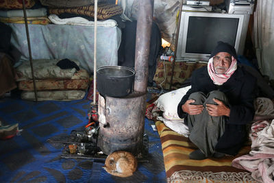 Old syrian refugee sits near a wood-burning fireplace in the cold inside a refugee tent.
