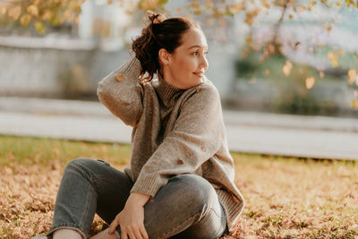 Woman looking away while sitting on grass and dry leaves