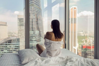 Rear portrait woman in white shirt sit on the bed window in a skyscraper near the bed