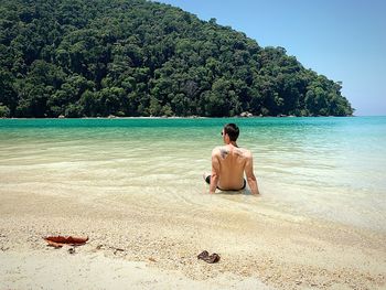 Rear view of shirtless young man sitting on beach against mountain during sunny day