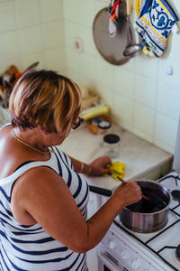 High angle view of mature woman pouring ingredient in saucepan on stove in kitchen 