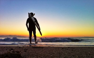 Silhouette man carrying surfboard on beach at dusk