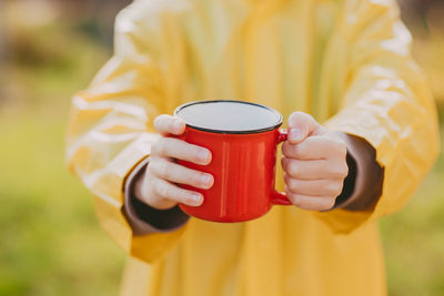 The hands of a boy in a yellow raincoat are holding hot cocoa in a red mug. cozy photo with a mug.