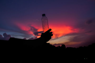 Silhouette hand holding plastic bottle against sky during sunset