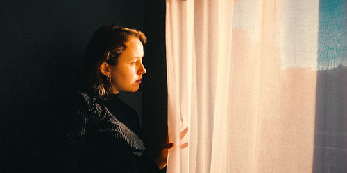 Side view of young woman looking through window at home