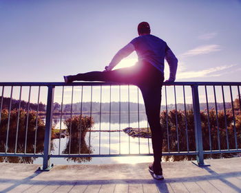 Man stretching legs before going run outside within sunny morning. outdoor exercising on lake bridge