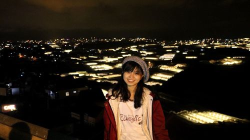 Portrait of smiling young woman standing against illuminated cityscape at night