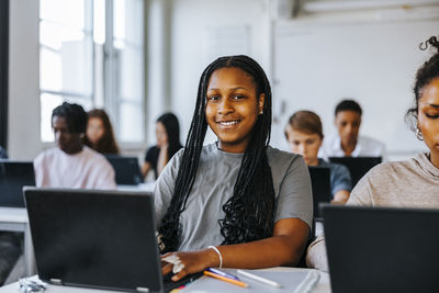 Portrait of teenage student with braided hair sitting with laptop in classroom at junior high school