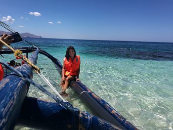 Portrait of young woman wearing life jacket sitting on boat in sea against blue sky