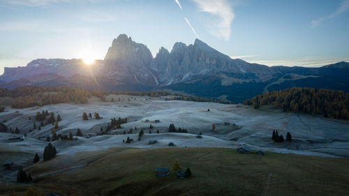 Sun rising above high alpine pasture with huts from behind a wall of mountains, aerial, dolomites