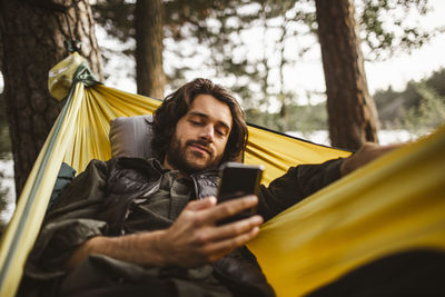 Young man using smart phone while lying over hammock in forest