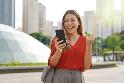 Brazilian surprised girl watching joyful her smartphone with sustainable city on the background