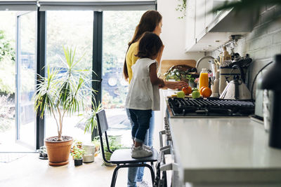 Girl standing on chair by woman preparing food in kitchen