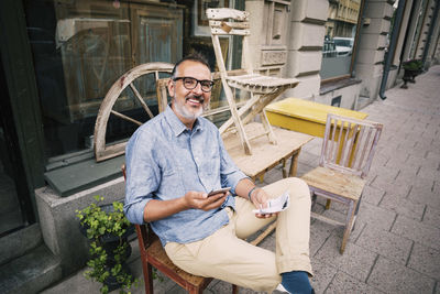 Portrait of owner holding mobile phone while sitting outside antique shop