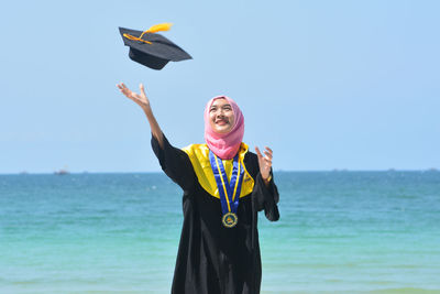 Woman in hijab looking at mortarboard while standing against sea