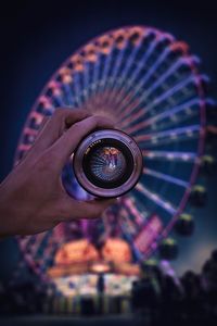 Cropped hand holding camera against illuminated ferris wheel at night