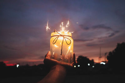 Cropped hand holding burning sparklers in container against sky at night