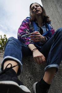 Low angle portrait of young woman sitting on retaining wall against sky