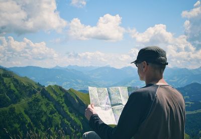 Rear view of man holding map with mountains in background against sky