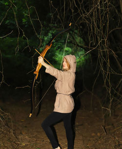 Young woman holding bow and arrow while standing in forest