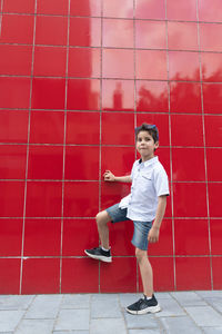 A cute boy in a white shirt raises his leg against a red wall, the football