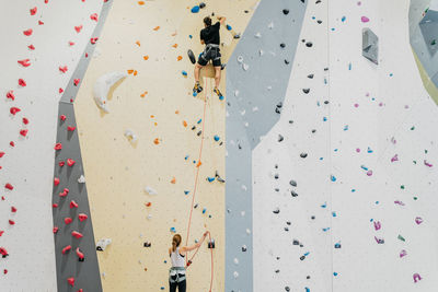 Back view of anonymous woman in activewear helping anonymous male alpinist to climb artificial wall during training in modern gym