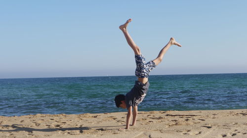 Full length of boy doing handstand at beach against clear sky during sunny day