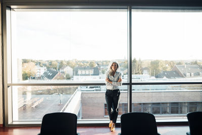 Businesswoman standing with arms crossed in front of glass window at work place