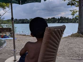 Rear view of shirtless boy sitting on chair at beach against sky