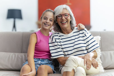 Happy girl sitting with arm around by grandmother on sofa at home
