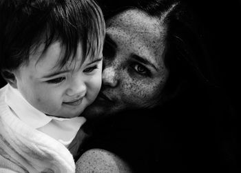 Portrait of smiling mother kissing cute baby son against black background