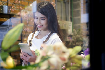 Smiling young woman using mobile phone at store seen through window