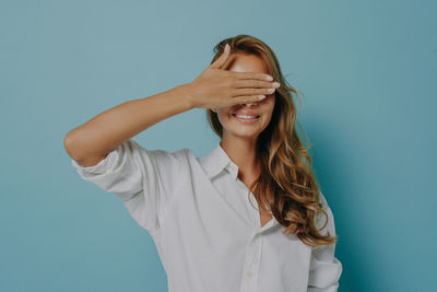 Smiling woman covering eyes standing against wall