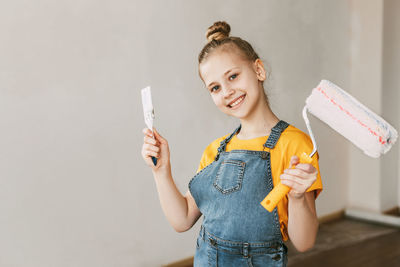 A girl in a denim overalls and a yellow t-shirt helps to paint the walls in an apartment white.