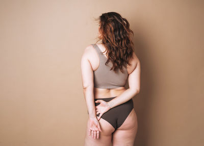 Young attractive woman plus size in underwear from back on beige background, body love