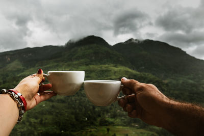 Cropped hands of couple toasting coffee cups against mountain