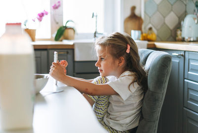 Little girl with long hair in home clothes using mobile at dinning table in kitchen at home