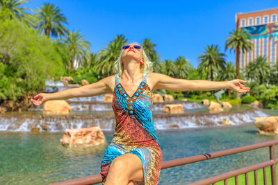 Smiling woman with arms outstretched sitting on railing with fountain in background