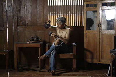 Shirtless man pouring tea in cup while sitting on chair at home