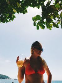 Woman wearing bikini holding drink while standing at beach against sky