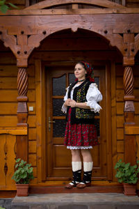 Thoughtful young woman in romanian clothing standing on porch of wooden house