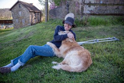 Smiling young man and his old dog cuddle in grass on rural farm hill with crutches