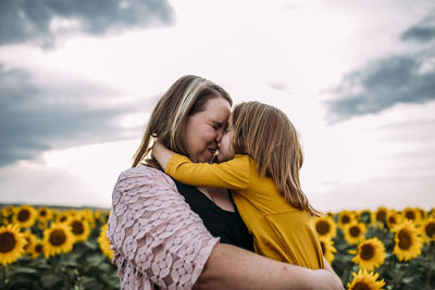 Mother and daughter embracing while standing in sunflower farm against sky