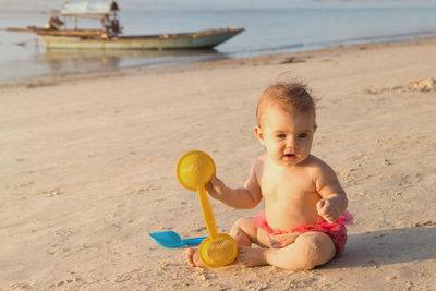 Cute little baby girl is sitting on a sandy beach near to sea and playing with plastic toys.