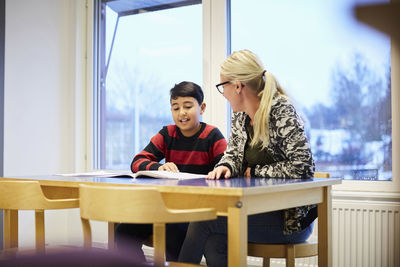 Mid adult teacher looking at boy reading book in classroom