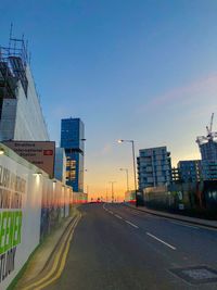 Road by buildings against sky during sunset