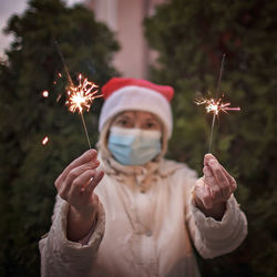 Portrait of senior woman wearing mask holding sparklers against trees